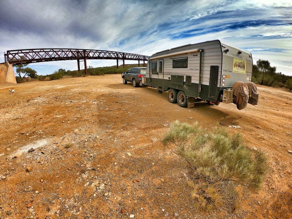 Car and caravan on a dusty road, near a railroad birdge underpass.