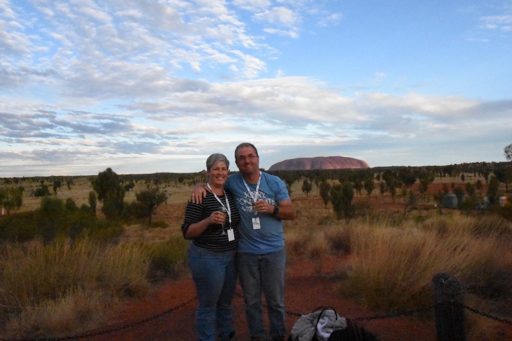 Man and woman standing in front of Uluru / Ayers Rock, holding a glass of wine