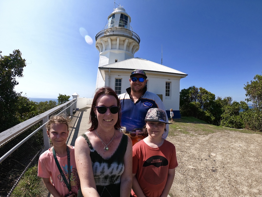 The Rig Family (mum, dad and two children) in front of a white lighthouse with blue sky in the background