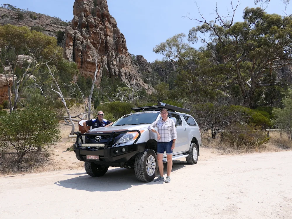 Couple standing either side of a silver ute, on sandy ground.