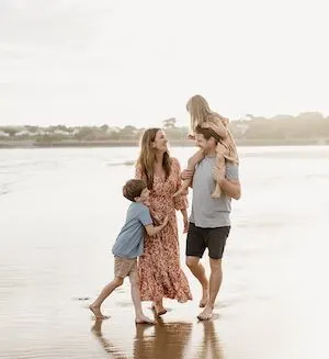 Smiling family of four on a beach, dad has daughter on his shoulders and the boy has his arms wrapped around his mum.