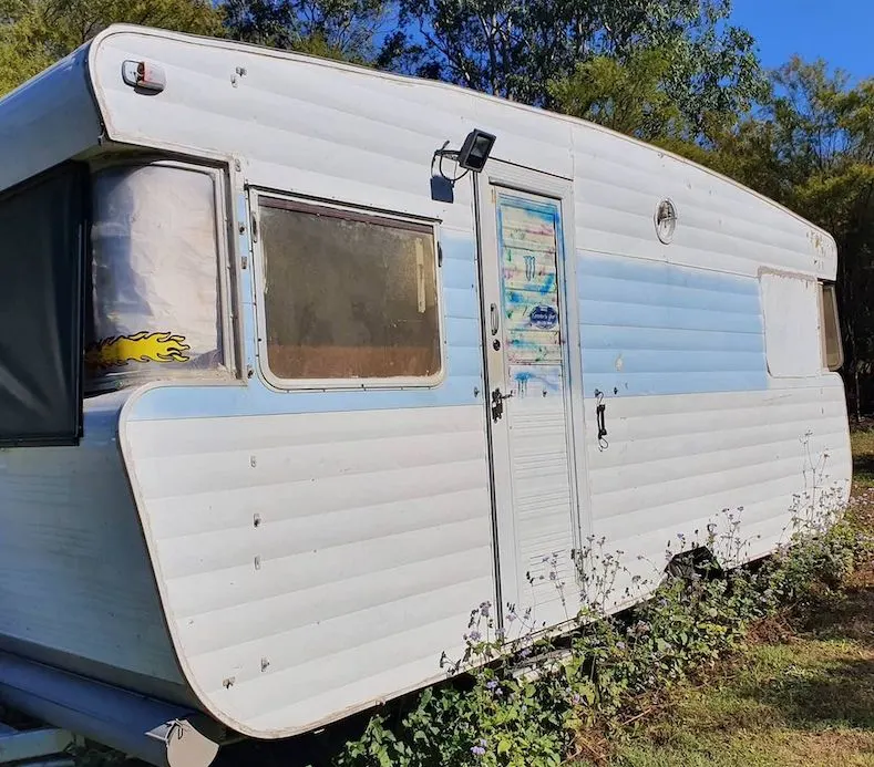 Old caravan in disrepair with grass growing up around it.