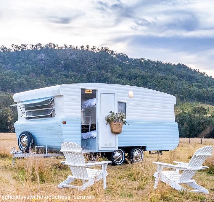 Renovated blue and white vintage caravan in a field with white lawn chairs arranged in front.